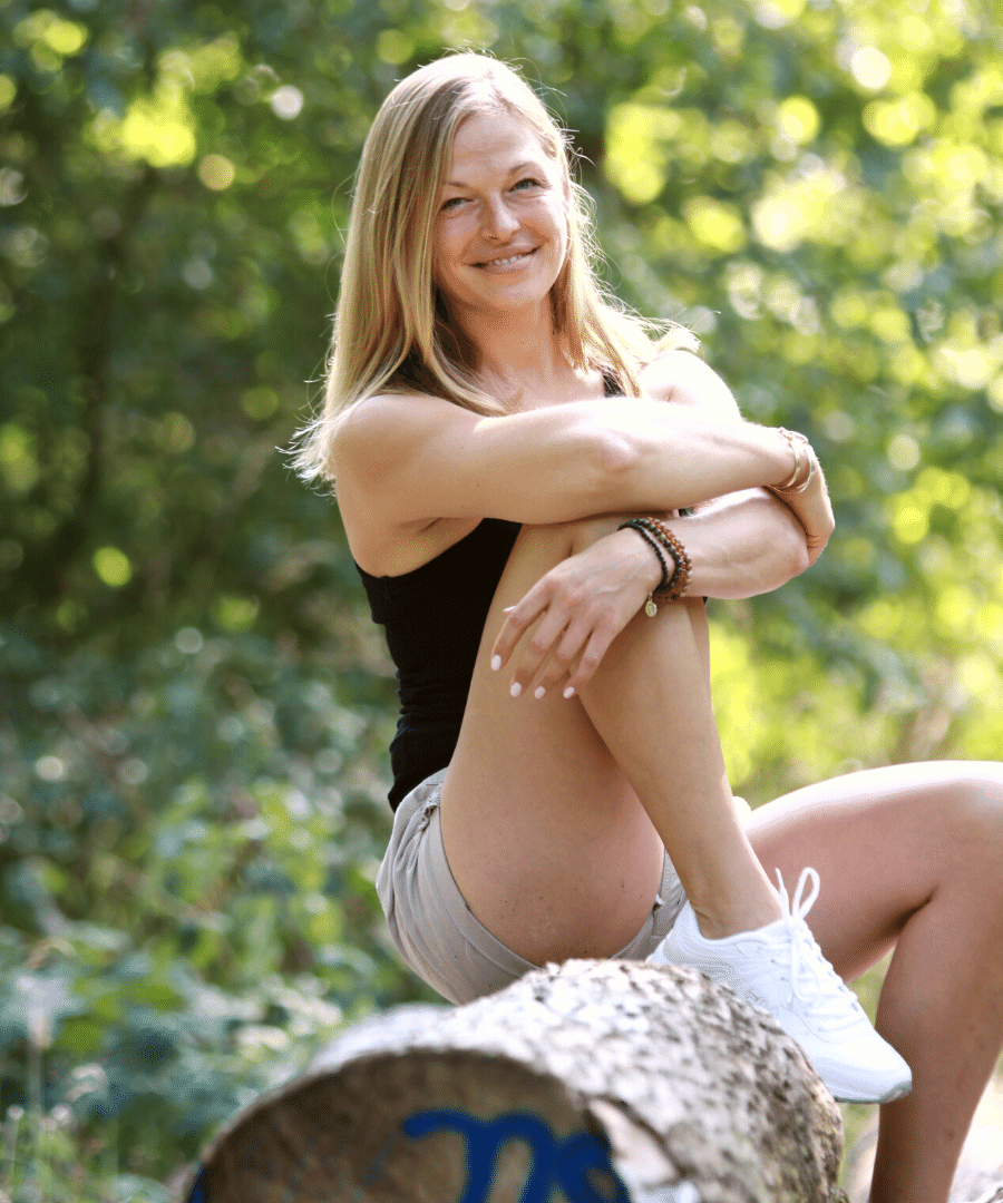 Blonde-woman-sits-in-the-forest-on-felled-tree-trunk-with-one-leg-bent-and-smiles-relaxed-and-friendly-into-the-camera