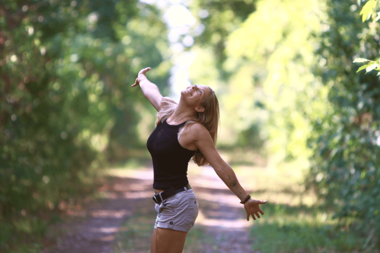 Blonde-woman-on-forest-path-dances-with-outstretched-arms-smiling-in-the-sunshine