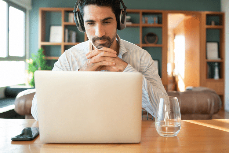 Frontal-shot-of-black-haired-man-between-40-and-50-with-headphones-in-living-room-sitting-at-dining-table-with-folded-hands-and-looking-interested-and-actively-listening-on-laptop