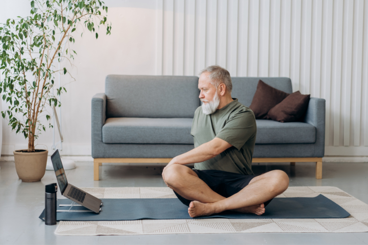 elderly-gentleman-sitting-cross-legged-in-front-of-PC-on-yoga-mat-in-living-room-while-easy twisting