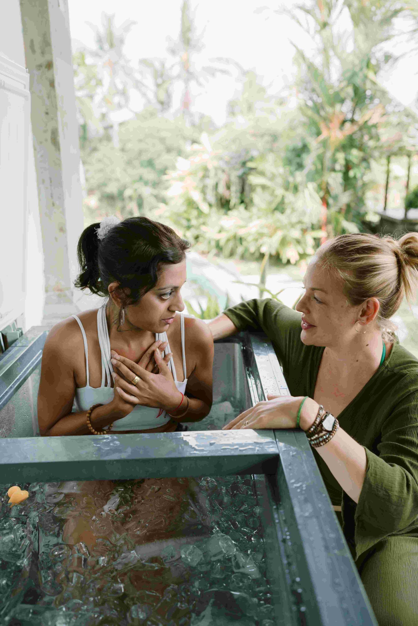 Woman-with-black-hair-indian-woman-in-icebath-wearing-white-bathingsuite-receives-support-by-female-facillitator-blonde-with-green-dress