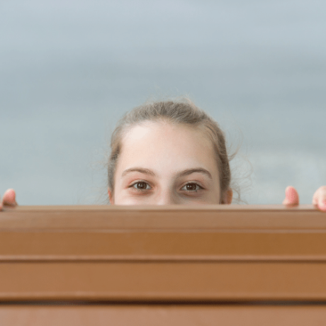 Was meine Arbeit ausmacht_Neugier_Mädchen schaut über Holzpaletten Girl-peeks-cheekily-and-happily-out-of-hiding-behind-wooden-wall-so-you-can-only-see-head-and-eyes